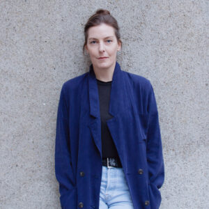 Irish Writer Caoilinn Hughes leans against a stone wall. She wears her dark hair pulled back, light jeans, and a dark blue baggy jacket.