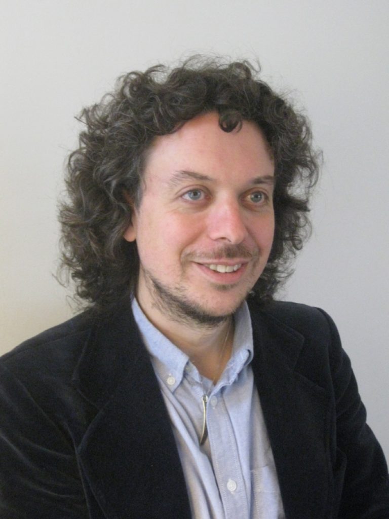 laurence cox with curly brown hair and collar shirt and blazer