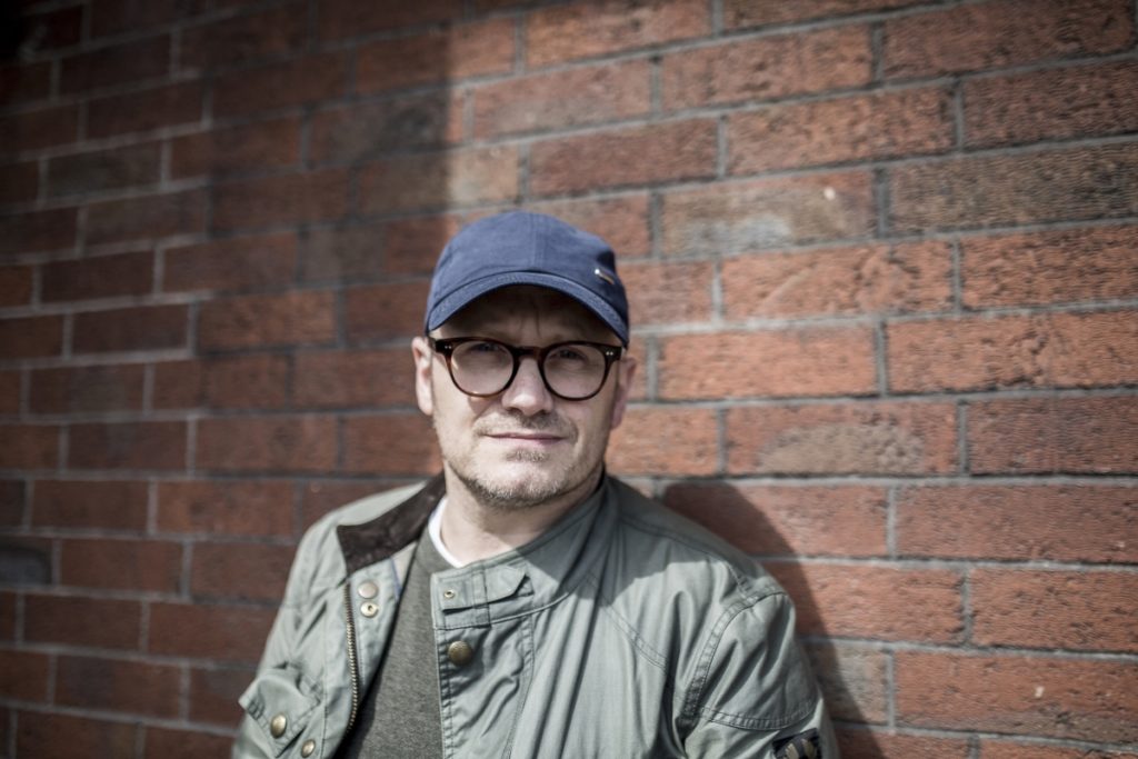 lenny abrahamson stands by brick wall wearing navy baseball hat and glasses